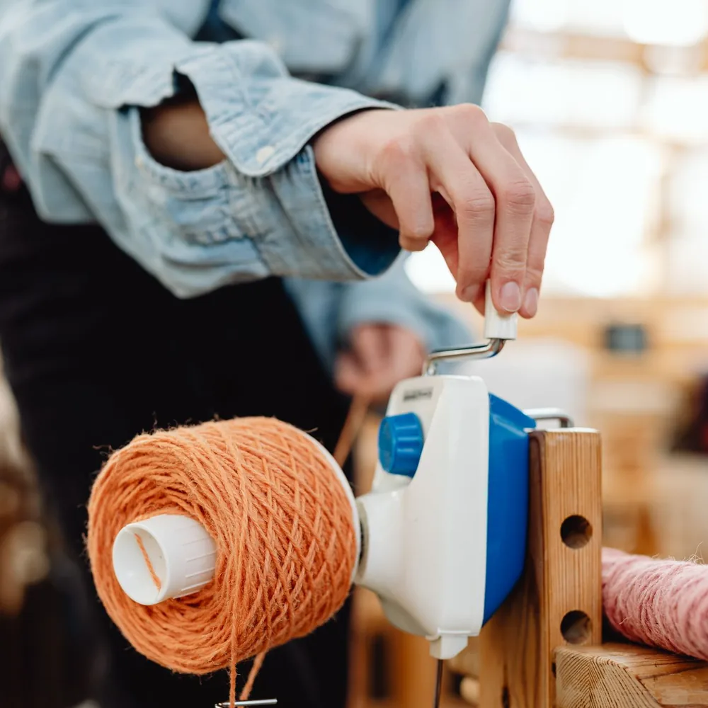 Cono de hilo naranja montado en un soporte de madera dentro de un taller textil, con las manos preparando la hebra antes del mechado.